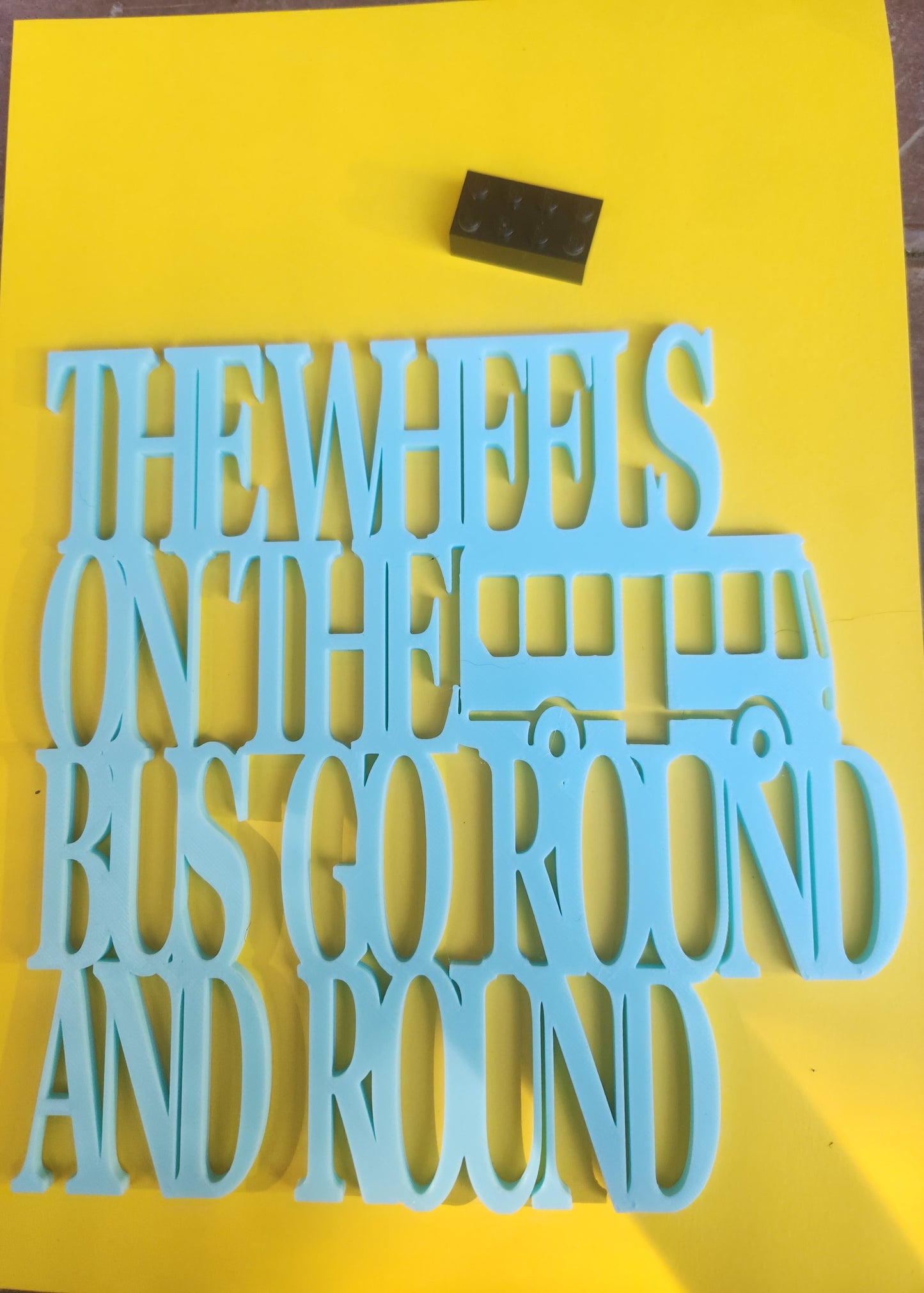 The Wheels on the Bus Go Round and Round 3D printed sky blue PLA wall art sign with bus design, shown flat on yellow background with Lego brick for scale, 19cm x 19cm, made in NZ