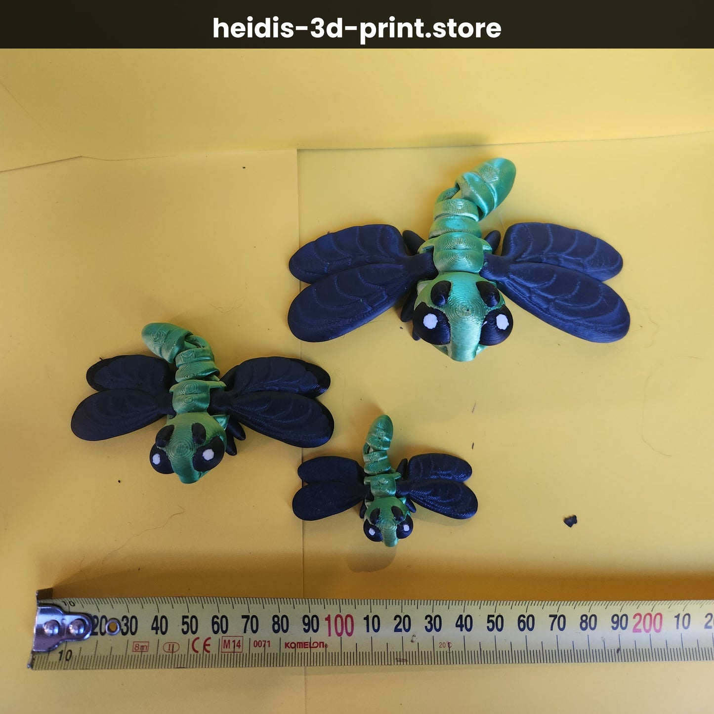 Three 3D printed dragonfly figures on yellow background next to ruler showing size comparison — small 60mm, medium 100mm, large 135mm wide, green and navy, Heidi's 3D Prints NZ
