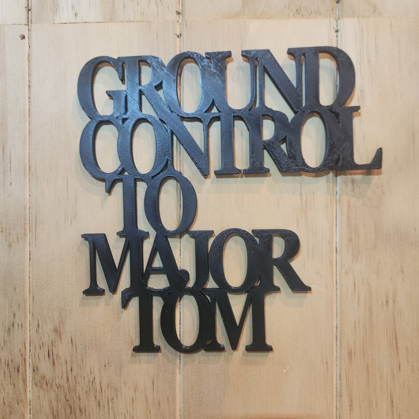 Ground Control to Major Tom 3D printed wall art sign in black, mounted on a warm wood-textured wall with natural light, showing the full 19cm × 19cm size and clarity of the lettering
