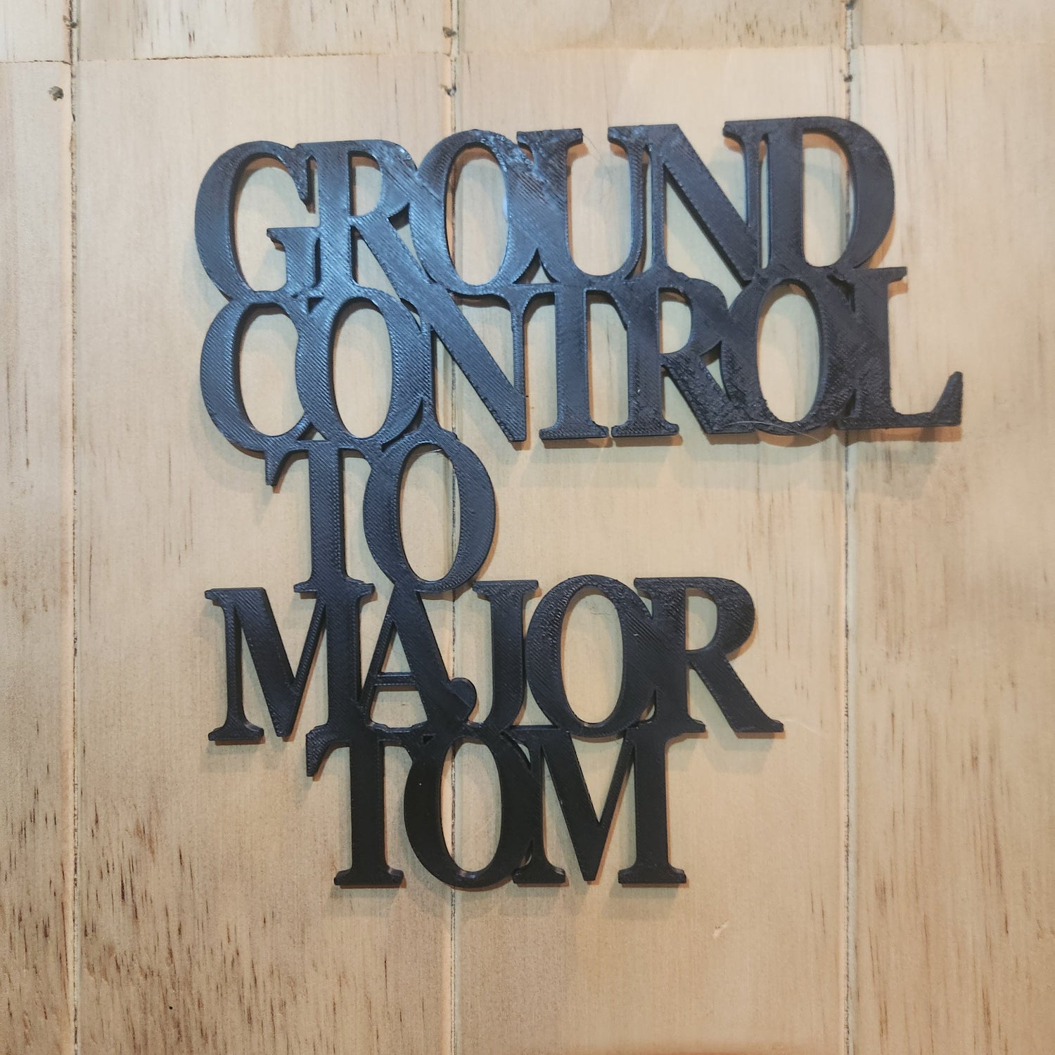 Ground Control to Major Tom 3D printed wall art sign in black, mounted on a warm wood-textured wall with natural light, showing the full 19cm × 19cm size and clarity of the lettering