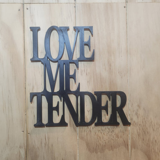 Love Me Tender 3D printed wall art sign in black on a wooden wall, showing the romantic phrase in bold 3D lettering