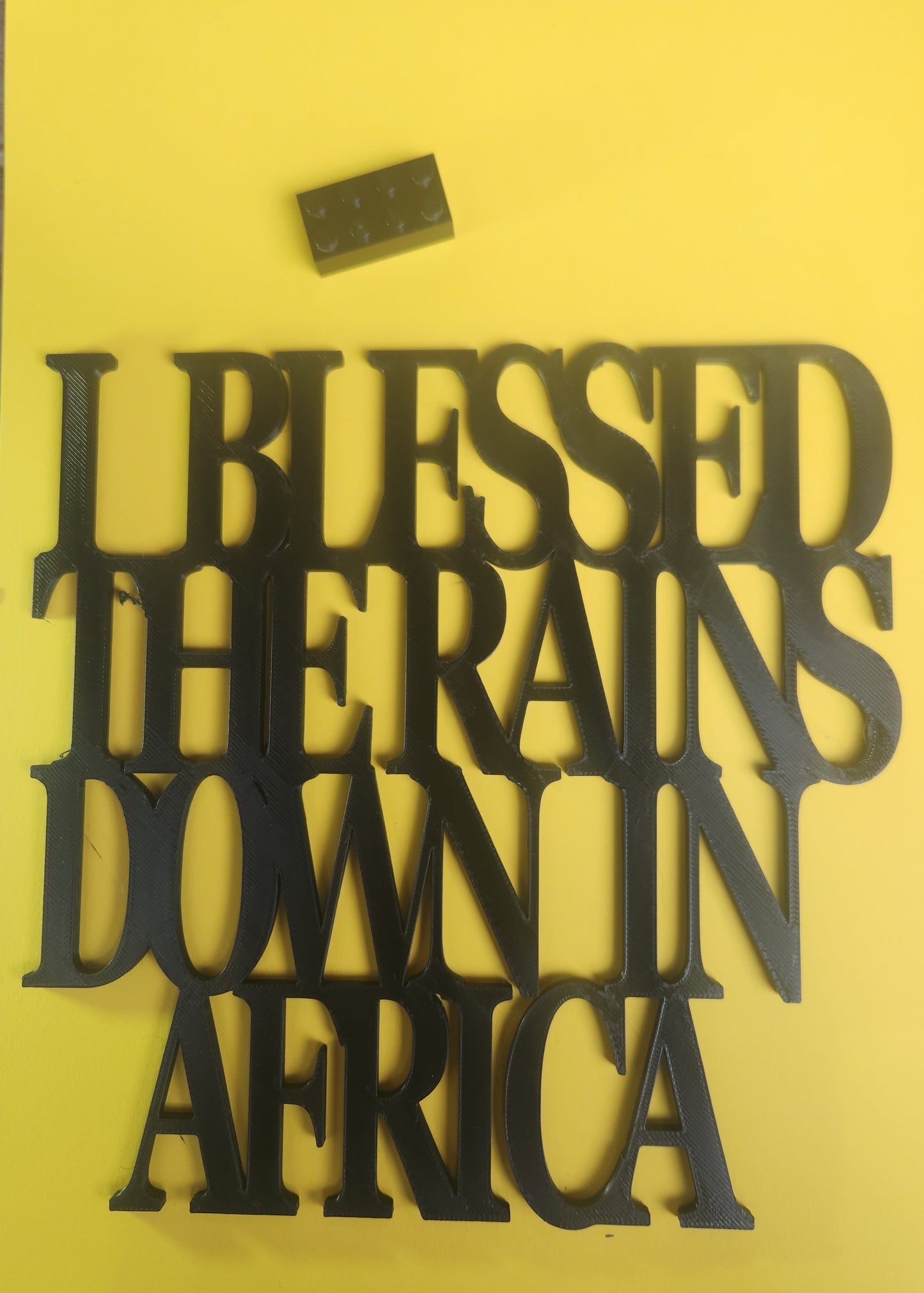 Close-up of I Blessed the Rains Down in Africa sign with Lego brick showing size reference, approximately 19cm × 19cm