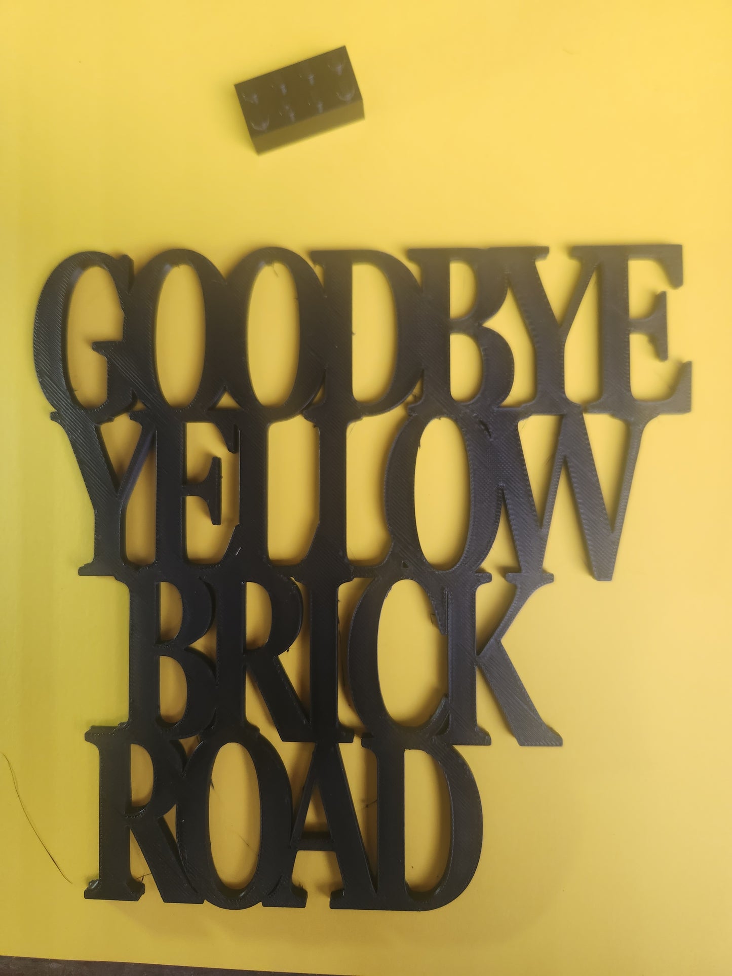 Goodbye Yellow Brick Road 3D printed black PLA wall art sign shown flat on a yellow background with a Lego brick for scale, bold stacked serif lettering, 19cm x 19cm, made in NZ