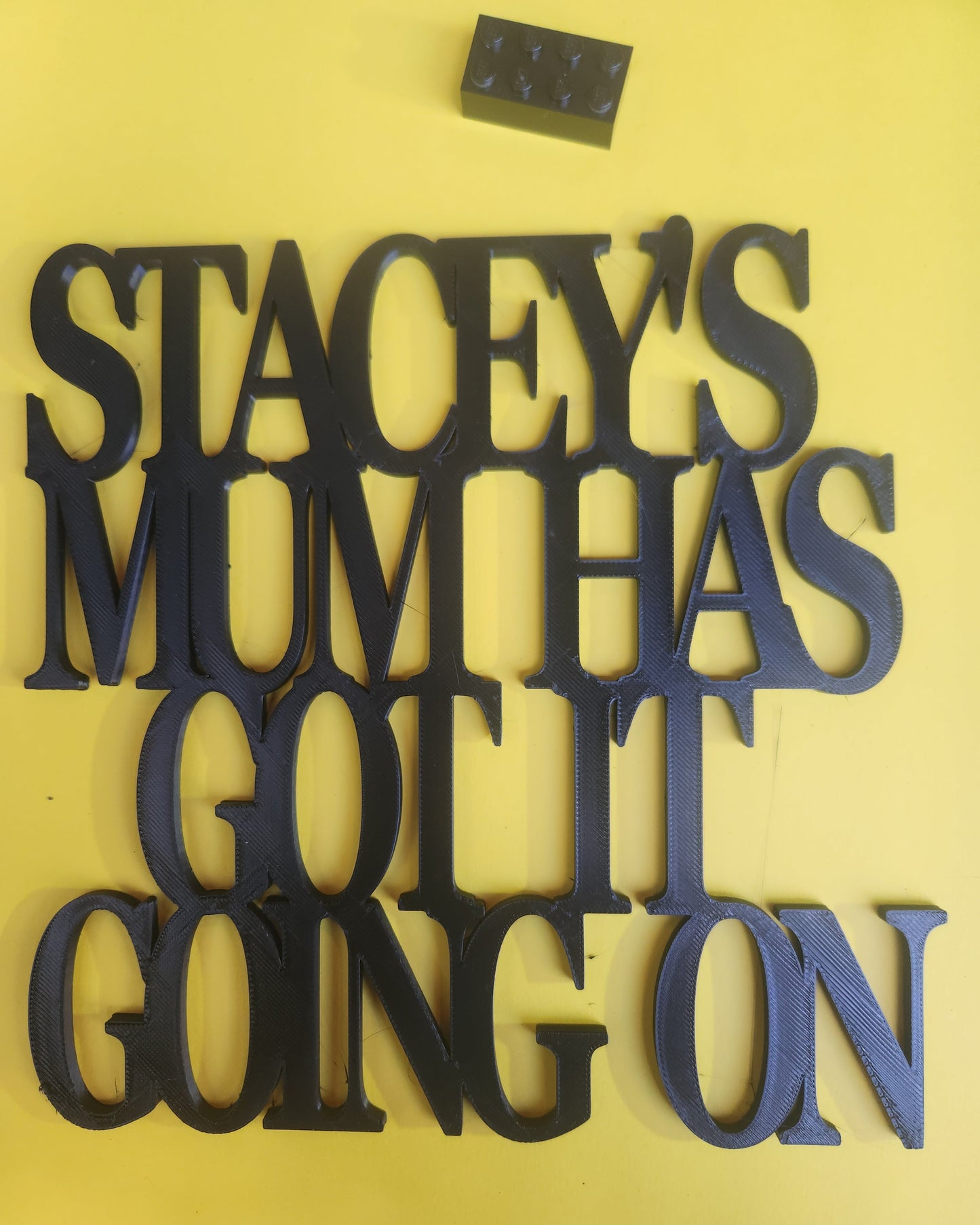 Stacey's Mum Has Got It Going On wall art sign in black PLA, photographed against a bright yellow background with a Lego brick placed beside it for scale