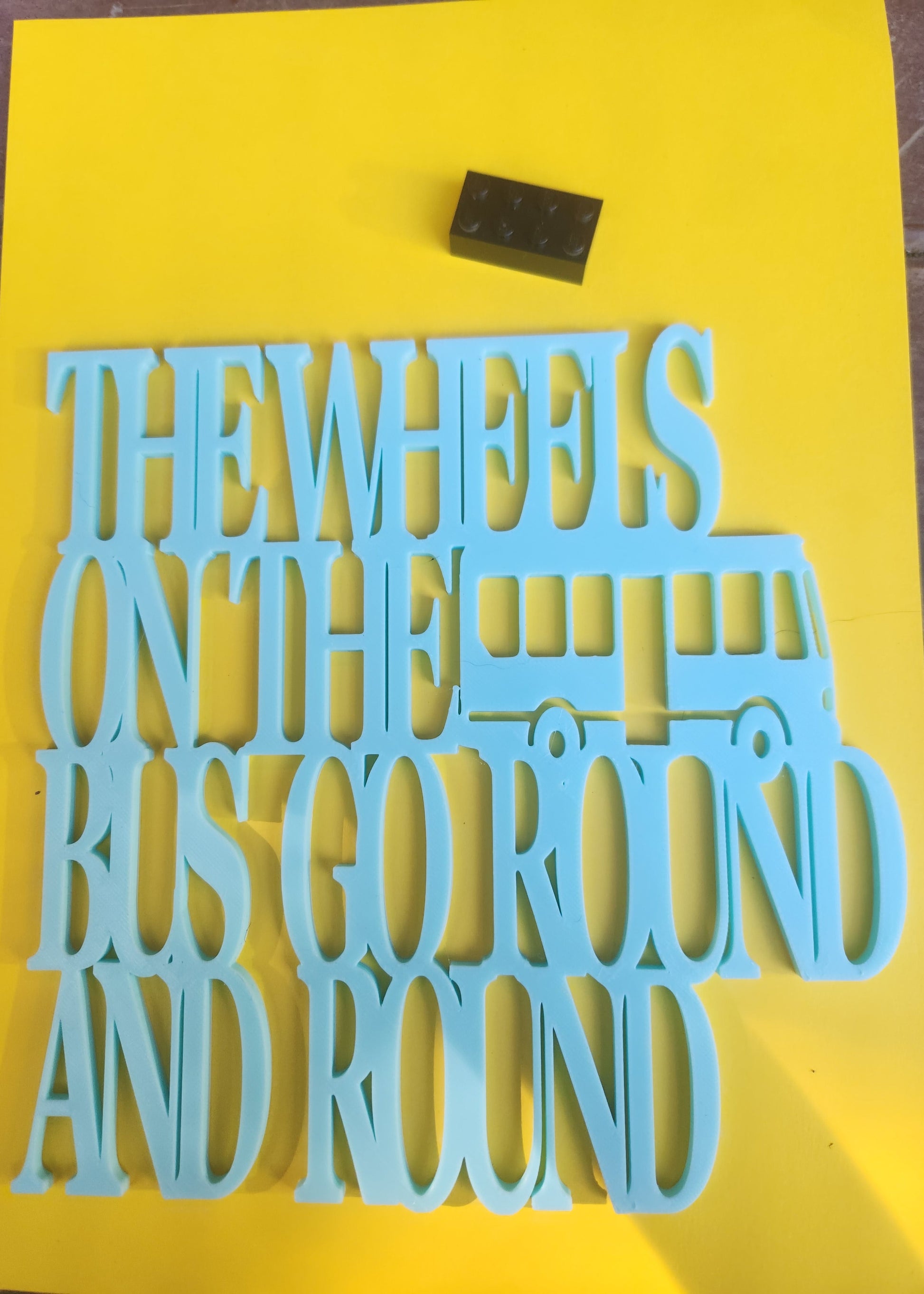 The Wheels on the Bus Go Round and Round 3D printed sky blue PLA wall art sign with bus design, shown flat on yellow background with Lego brick for scale, 19cm x 19cm, made in NZ