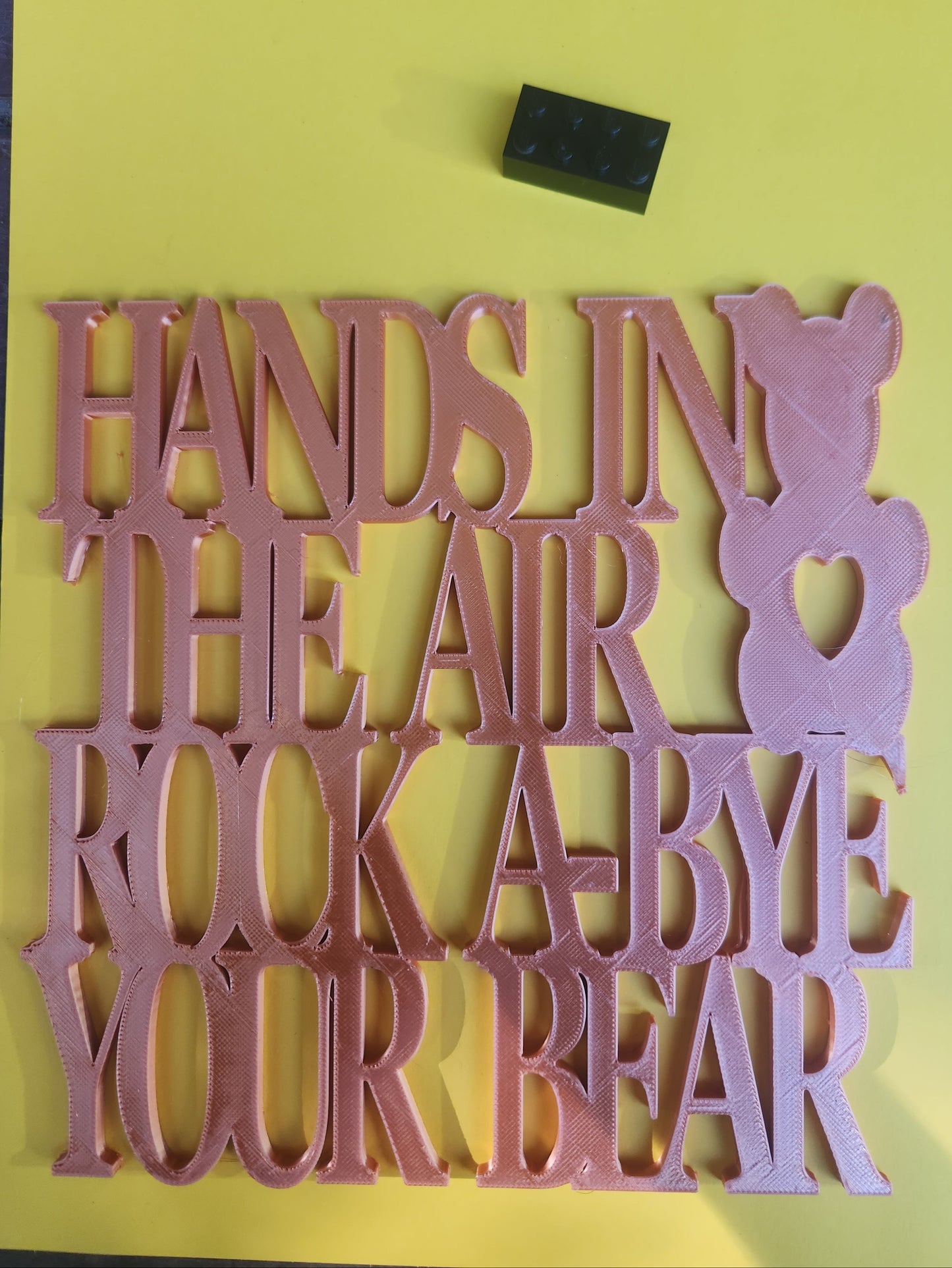 Hands in the Air Rock-A-Bye Your Bear 3D printed rose gold PLA wall art sign with teddy bear and heart design, shown flat on yellow background with Lego brick for scale, 19cm x 19cm, made in NZ