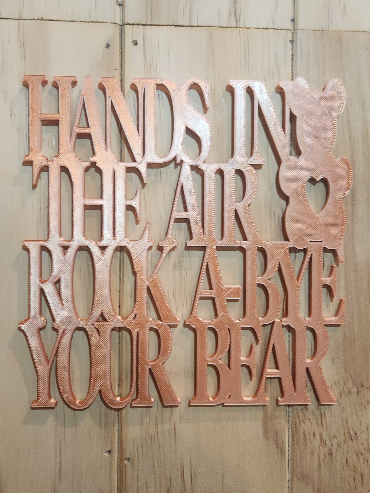 3D printed rose gold PLA wall art sign reading Hands in the Air Rock-A-Bye Your Bear with teddy bear silhouette, mounted on a whitewashed wood wall, 190mm x 190mm, handmade in Upper Hutt New Zealand