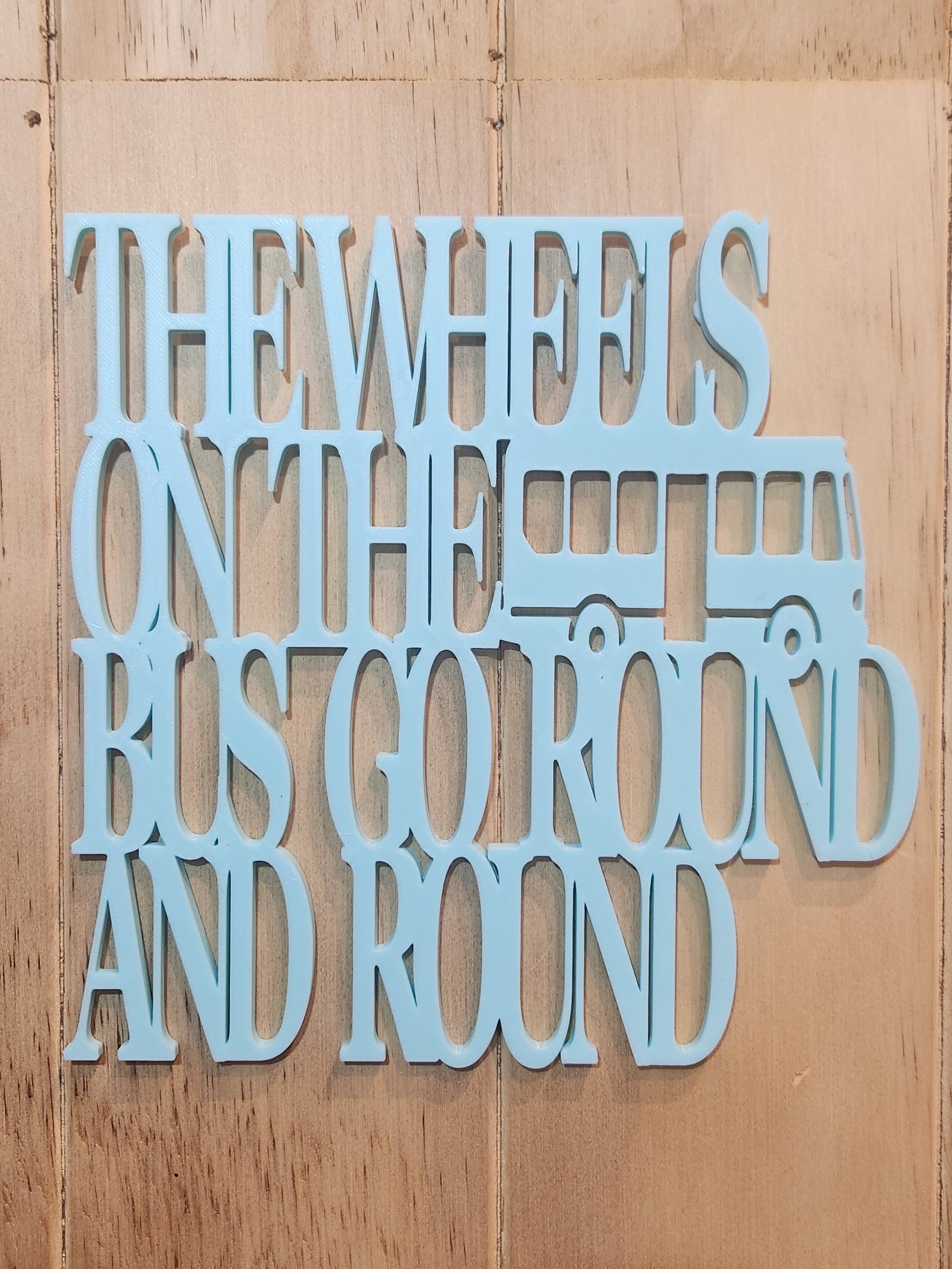 3D printed sky blue PLA wall art sign reading The Wheels on the Bus Go Round and Round with bus silhouette, mounted on a wood wall, 190mm x 190mm, handmade in Upper Hutt New Zealand