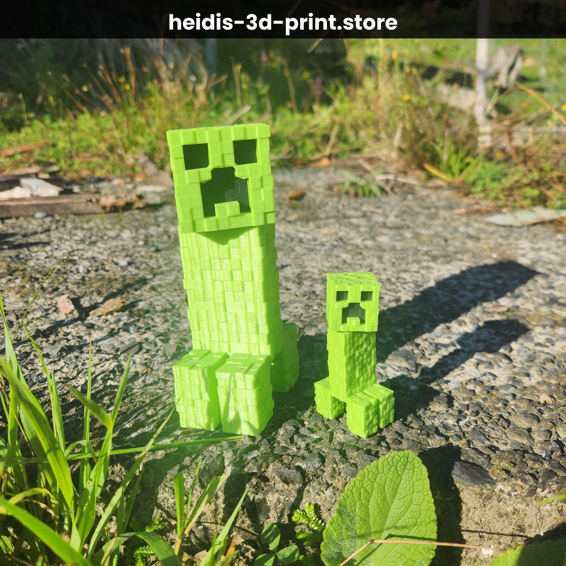 Large (150mm) and small (75mm) 3D printed Minecraft Creeper figures side by side on gravel, showing size comparison, green PLA, made in New Zealand by Heidi's 3D Prints