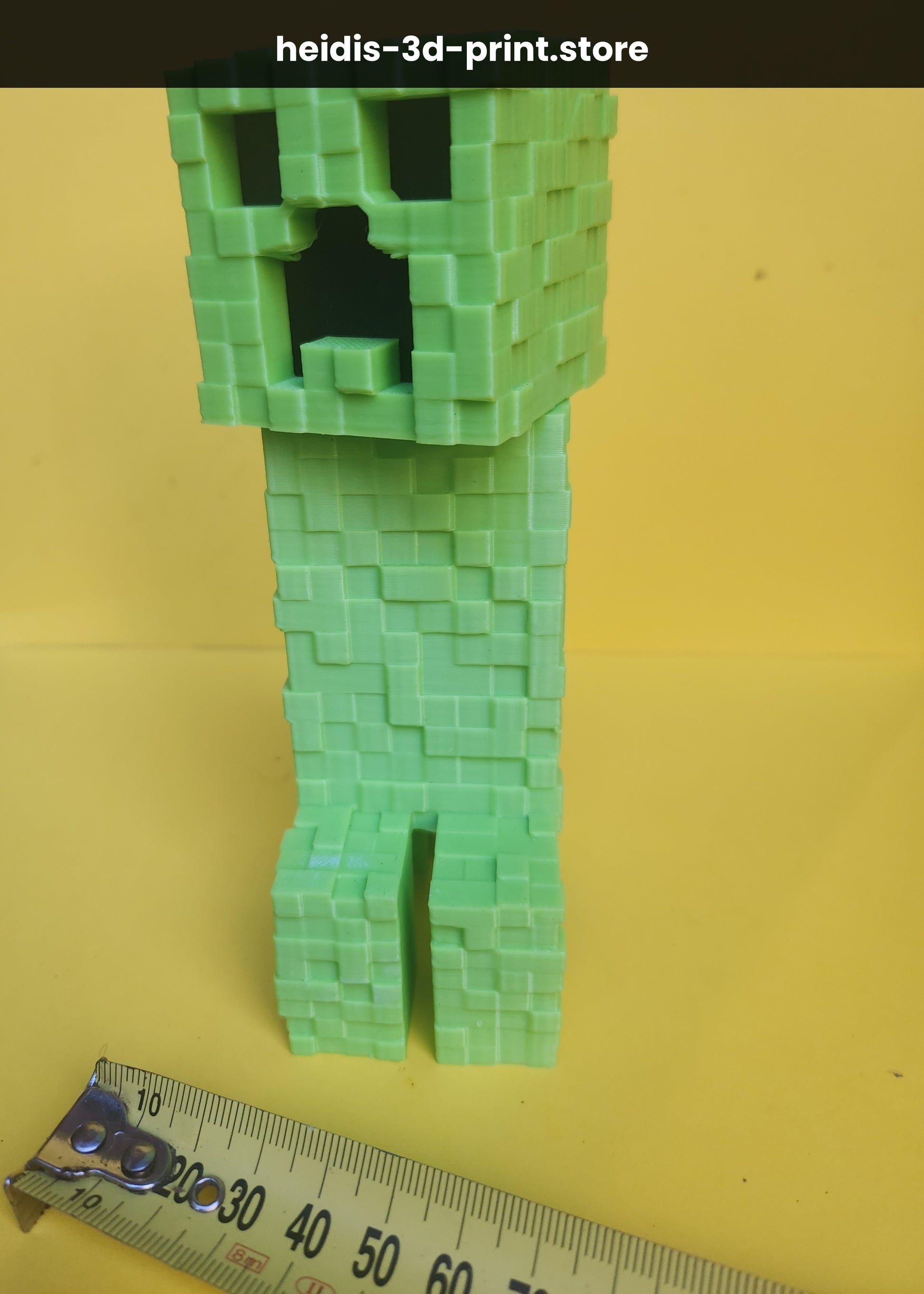 Angled view of small 3D printed Minecraft Creeper figure on yellow background with tape measure, showing pixel detail on blocky green body, made in NZ