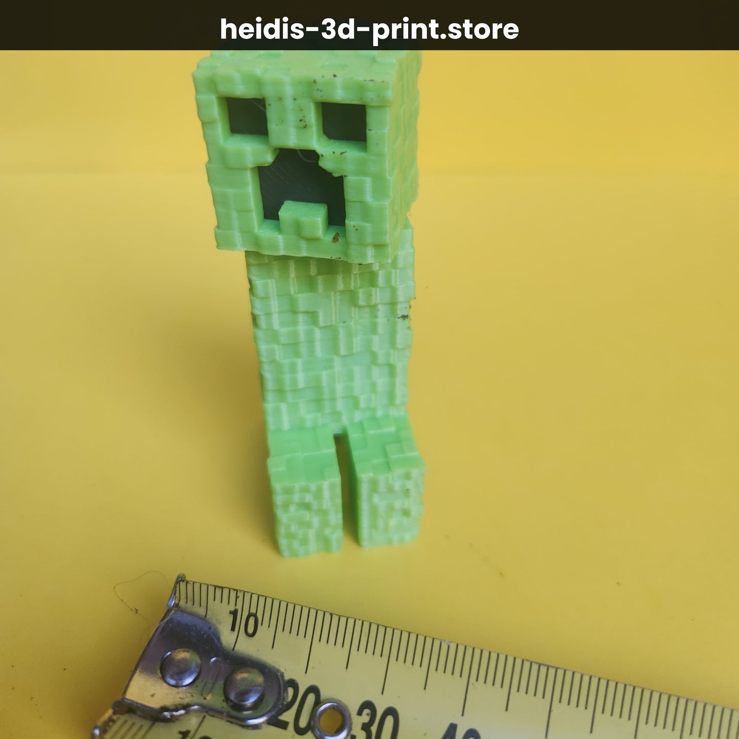 Small 3D printed Minecraft Creeper figure on yellow background next to ruler showing 75mm height, 23mm wide × 35mm long, green PLA, Heidi's 3D Prints NZ