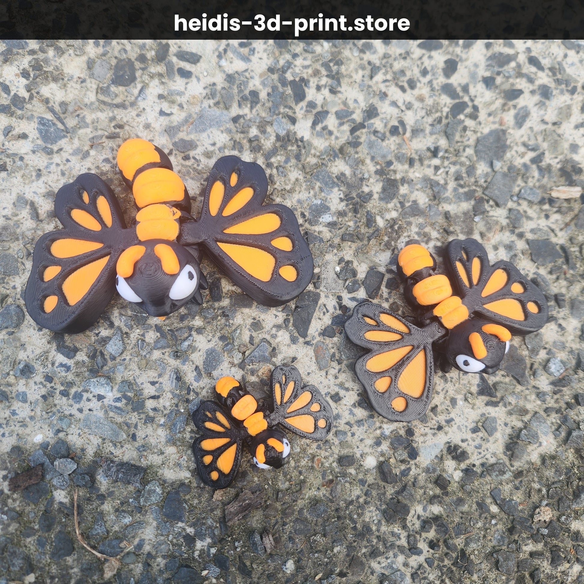 Overhead view of three 3D printed cute butterfly figures in three sizes on gravel, monarch butterfly wing pattern in orange and dark brown, made in Upper Hutt NZ