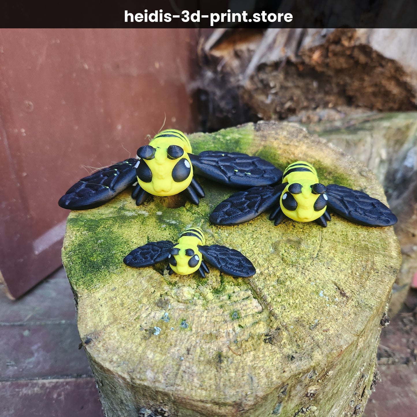 Three 3D printed articulated bee figures in small, medium, and large sizes sitting on a mossy tree stump, yellow and black PLA, handmade in Upper Hutt New Zealand
