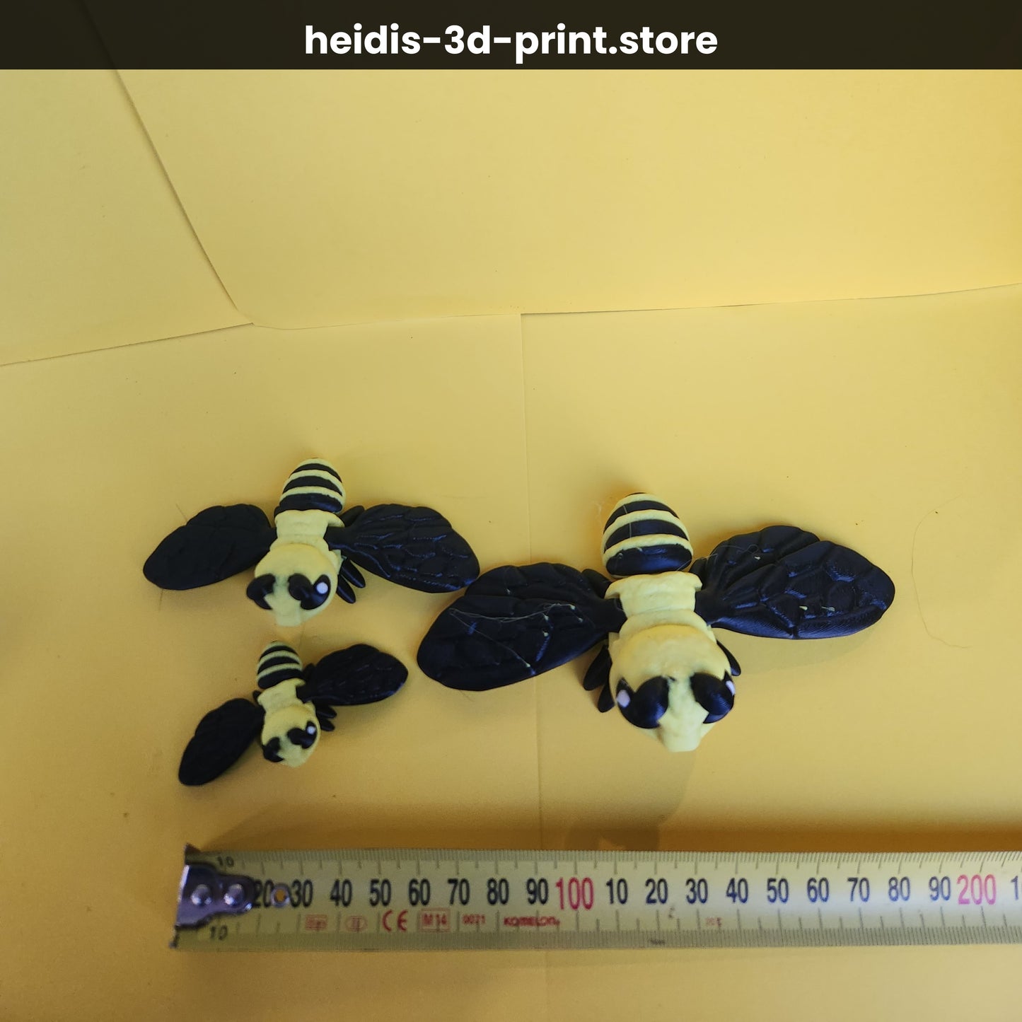 Three 3D printed bee figures on yellow background next to ruler showing size comparison, small ~55mm, medium 100mm, large 135mm wide, yellow and black, Heidi's 3D Prints NZ