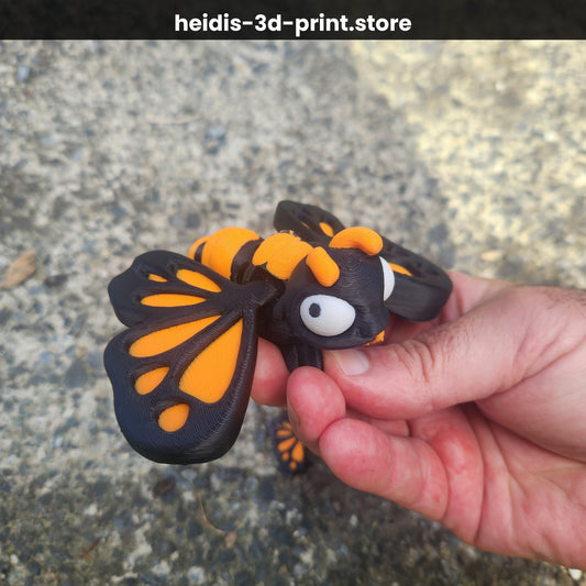 Hand holding a large 3D printed cute butterfly figure in monarch orange and dark brown, showing expressive white eyes and detailed wing pattern, made in Upper Hutt NZ by Heidi's 3D Prints