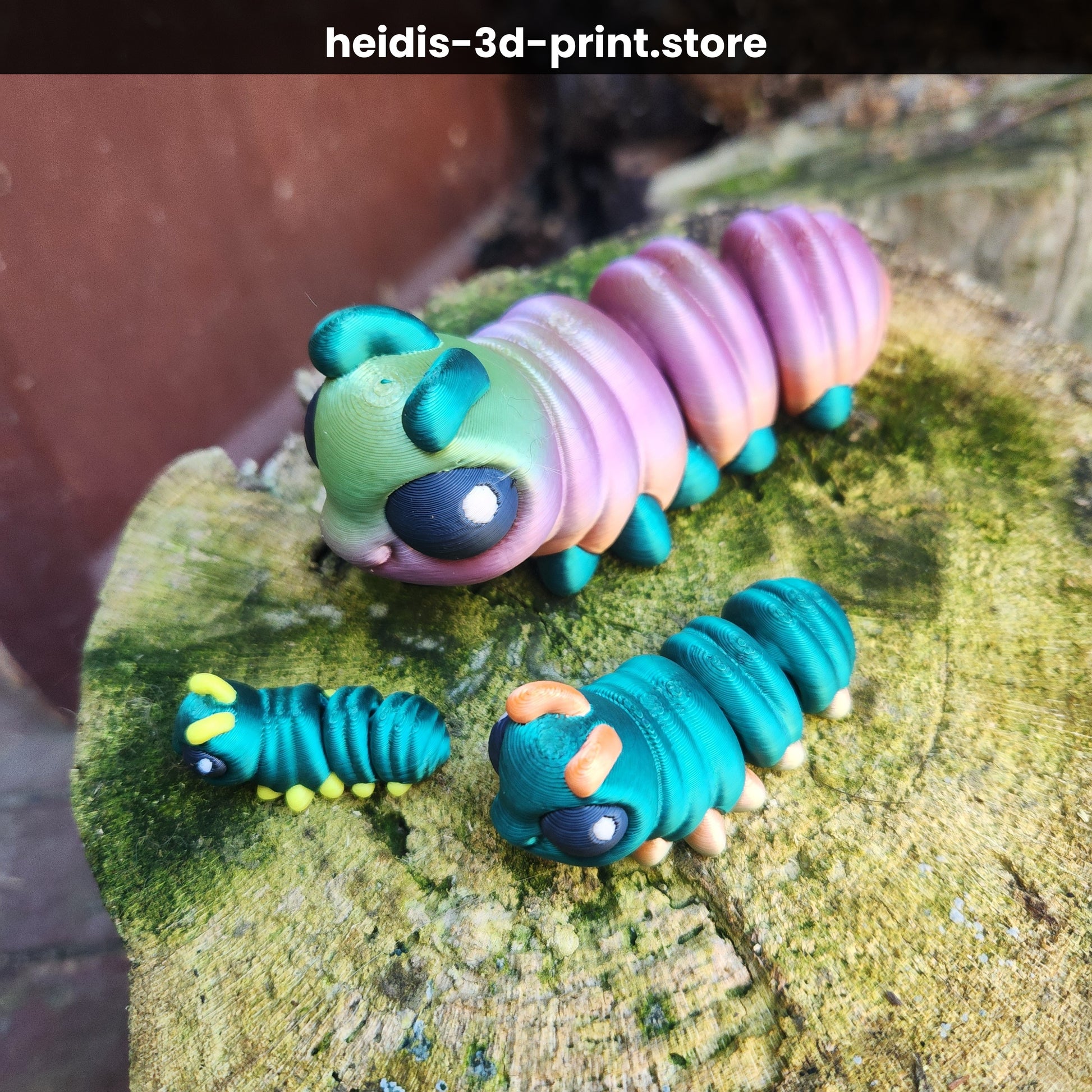 Three 3D printed articulated caterpillar figures in small, medium and large on a mossy tree stump — teal, pink/teal, and green colourways — handmade in Upper Hutt New Zealand