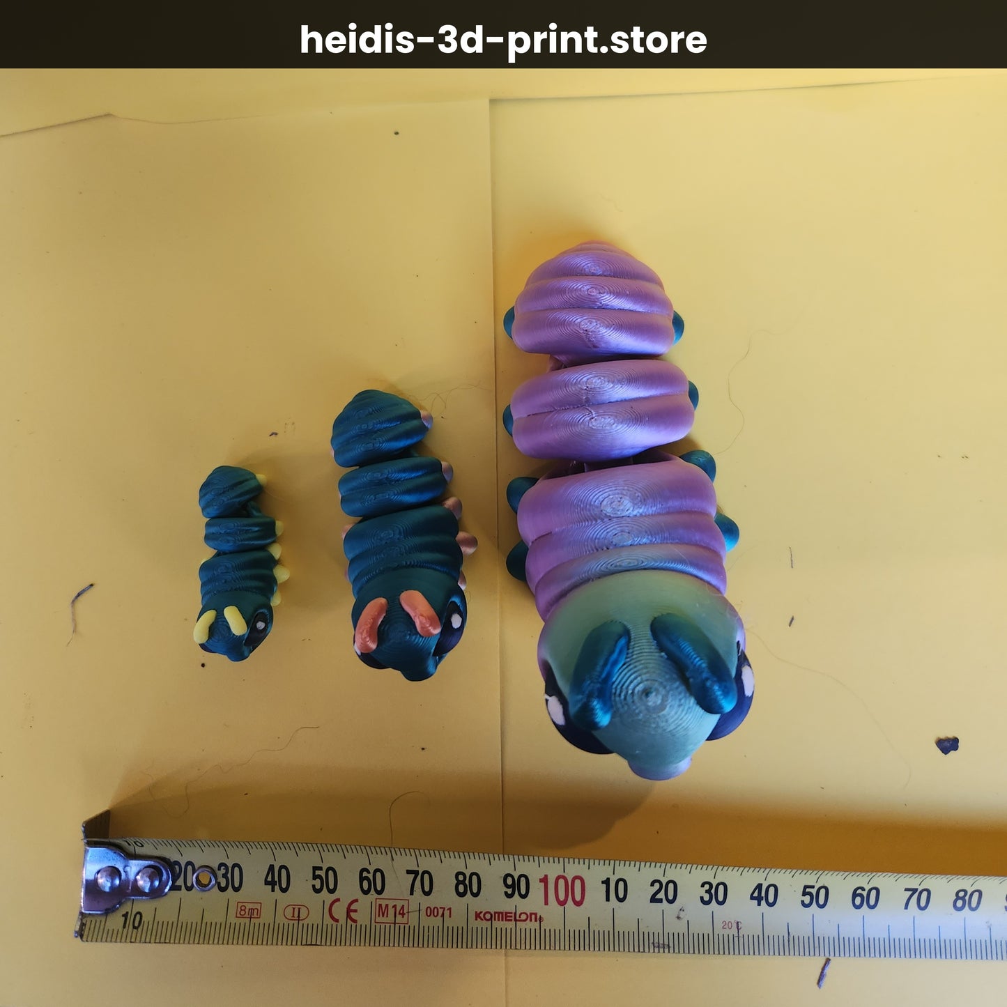 Three 3D printed caterpillar figures on yellow background with ruler: small 40mm long, medium 65mm long, large 105mm long — teal and purple colourways, Heidi's 3D Prints NZ