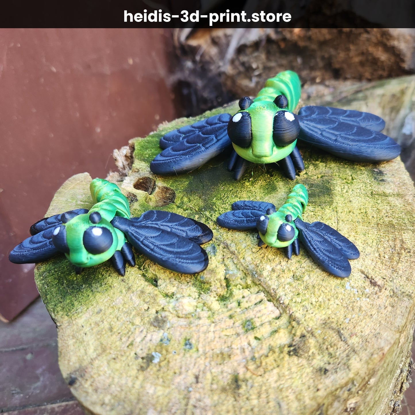 Three 3D printed articulated dragonfly figures in small, medium, and large sizes on a mossy tree stump, green and navy PLA, handmade in Upper Hutt New Zealand