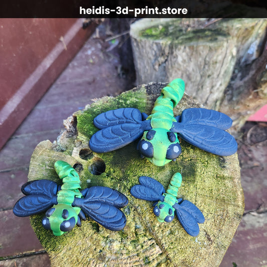 Three 3D printed dragonfly figures on a mossy stump showing face detail — large black eyes with white highlights, green articulated body, Heidi's 3D Prints NZ