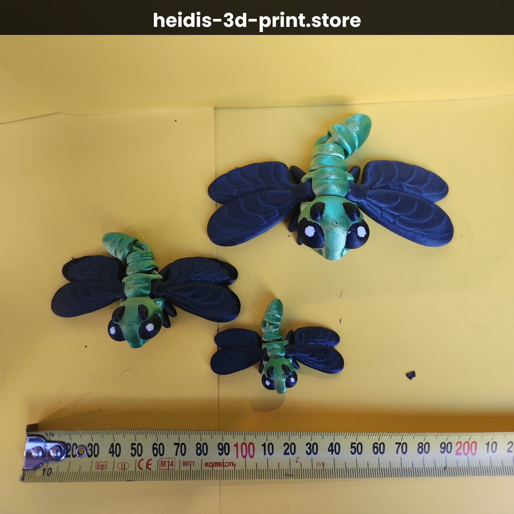 Three 3D printed dragonfly figures on yellow background next to ruler showing size comparison — small 60mm, medium 100mm, large 135mm wide, green and navy, Heidi's 3D Prints NZ