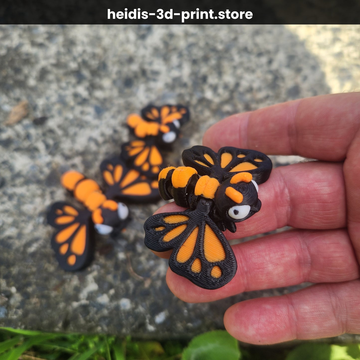 Hand holding small 3D printed cute butterfly figure with small butterfly visible in background on gravel, orange and dark brown monarch colours, NZ made