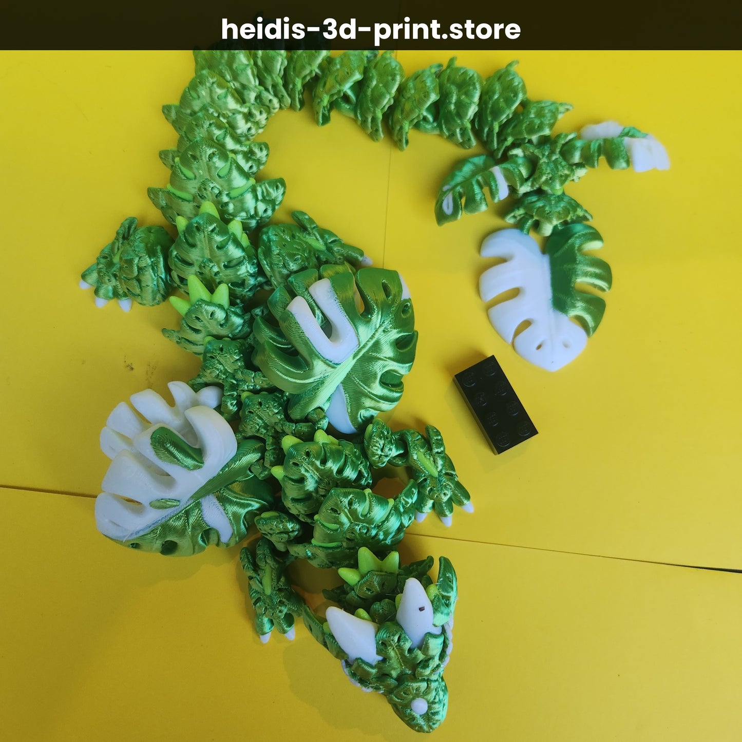 Overhead view of 3D printed Monstera Leaf Dragon on yellow background with Lego brick for scale — 470mm long, green articulated dragon figure, Heidi's 3D Prints NZ