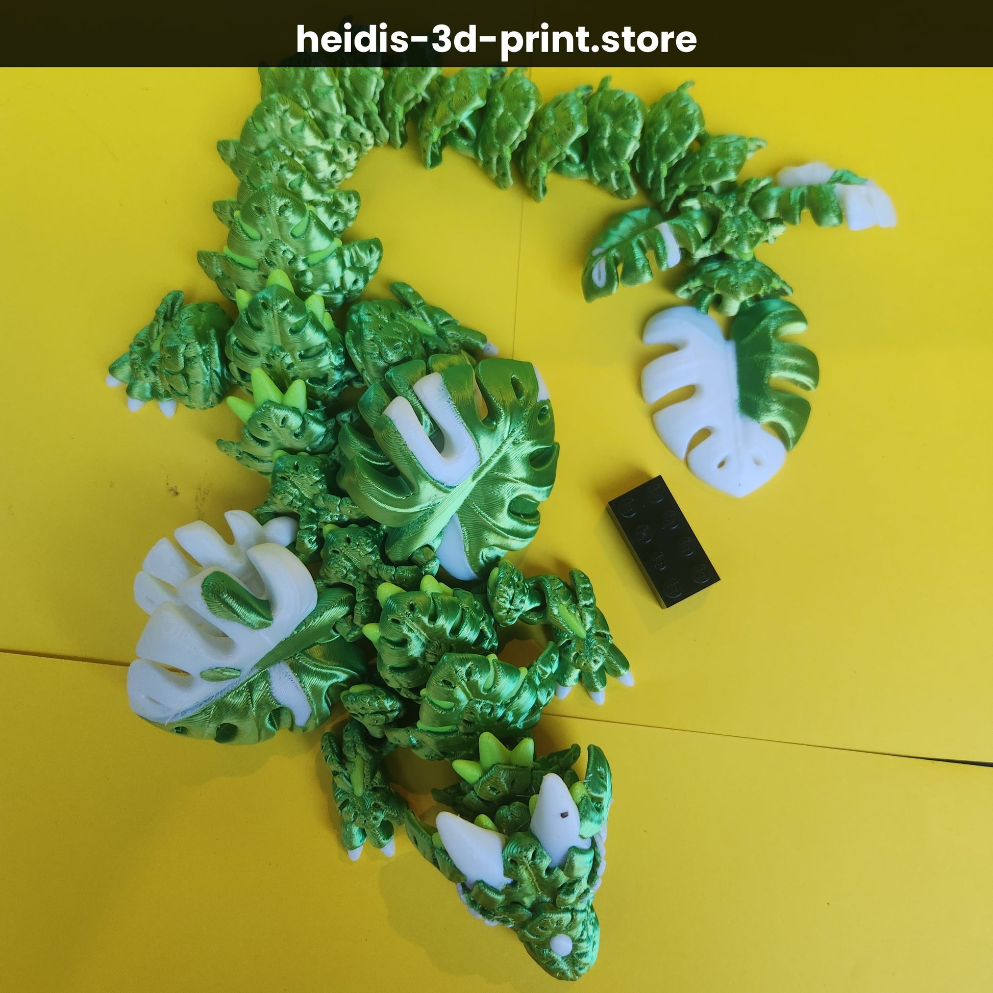 Overhead view of 3D printed Monstera Leaf Dragon on yellow background with Lego brick for scale — 470mm long, green articulated dragon figure, Heidi's 3D Prints NZ
