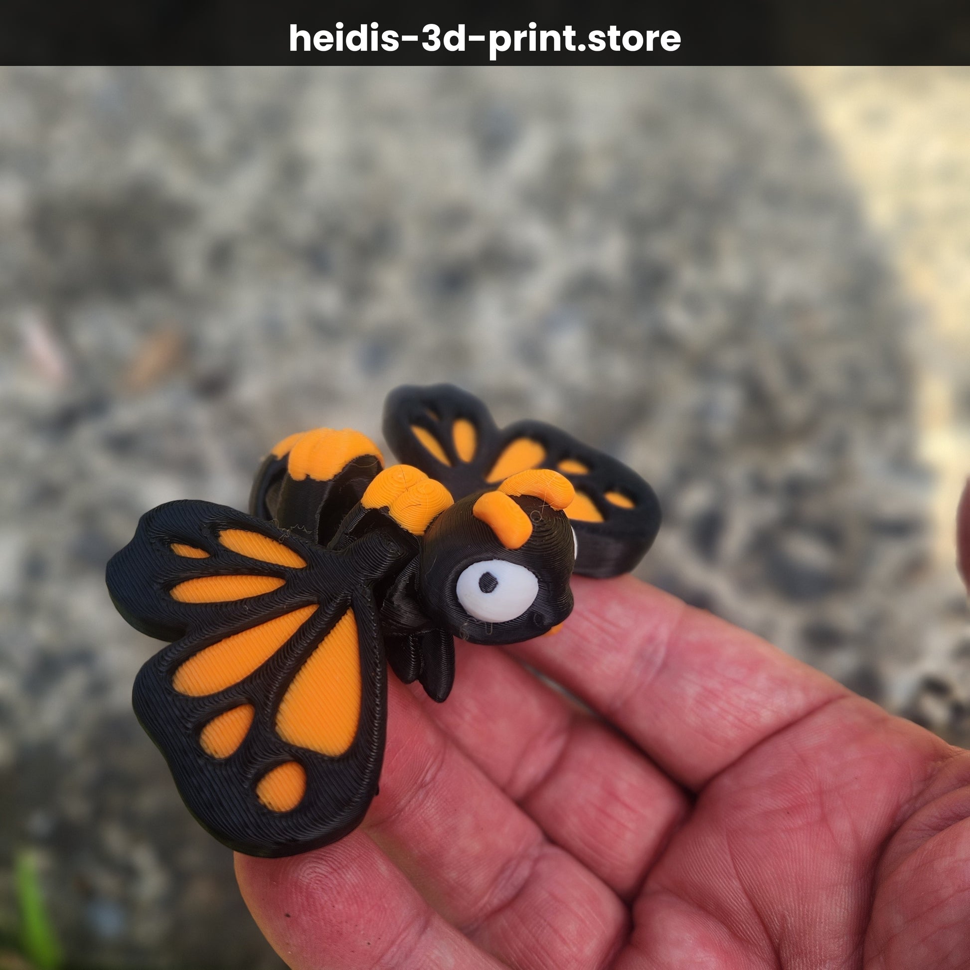 Hand holding medium 3D printed cute butterfly figure with small butterfly visible in background on gravel, orange and dark brown monarch colours, NZ made